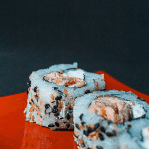 Un rollo de sushi con camarón, queso crema y jengibre. El camarón es rosado y brillante, el queso crema es blanco y cremoso, y el jengibre es naranja y picante.