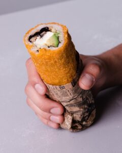 Una imagen de un Hand Roll de Carne + Queso Crema + Cebollín, envuelto en alga nori y panko crujiente. La carne es marrón y jugosa, el queso crema es blanco y cremoso, y el cebollín es verde y fresco. El alga nori es de color verde oscuro y el panko crujiente es de color dorado.